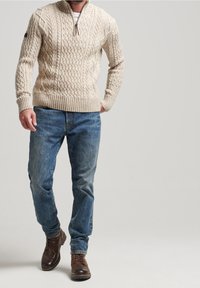 Beige Strickpullover mit hohem Kragen und durchgehendem Reißverschluss, der ein strukturiertes Kabelmuster aufweist. Getragen mit blauen Jeans und braunen Stiefeln.
