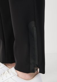 Zwarte sportbroek van gestructureerde stof, met een zijritsdetail en een glanzend zwart accent aan de zoom. Witte sneaker gedeeltelijk zichtbaar.