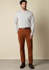 Šedý svetr s dlouhým rukávem, hnědé bavlněné kalhoty, hnědý kožený pásek, strukturované boty; stojí proti neutrálnímu pozadí.