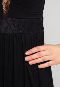Svart klänning med rynkad liv, spetsdetalj vid uringningen och dekorerad texturerad midja. Flödande kjol med plissédetaljer.