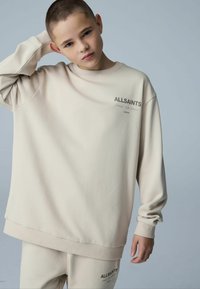 Sweat-shirt ample de couleur beige clair avec poignets et ourlet côtelés. Il présente le logo "ALLSAINTS" imprimé en foncé sur le devant.
