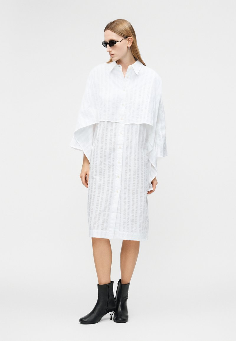 Femme portant une robe midi blanche boutonnée avec des manches larges superposées, des bottines noires et des lunettes de soleil noires, se tenant devant un fond uni.