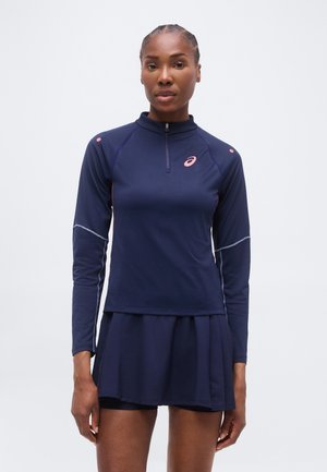 Femeie îmbrăcată într-un top sportiv cu mâneci lungi, albastru închis, cu fermoar și o fustă plisată asortată, stând în fața unui fundal simplu.