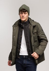 Oliwkowa kurtka z odpinanym czarnym pikowanym podszewką, dużym kołnierzem, kieszeniami z przodu i zapięciem na guziki, noszona na białym t-shircie.