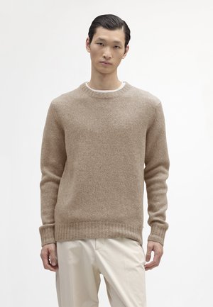 Beige stickad tröja med rund halsringning och ribbade ärmar, gjord av mjukt, texturerat tyg. Kombineras med ljusa byxor.