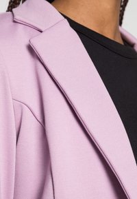 Vaaleanliila bleiseri, jossa on rakenteellinen kaulus, valmistettu pehmeästä kankaasta, yhdistetty mustan t-paidan kanssa alla. Korostuu sileä rakenne ja istuva malli.