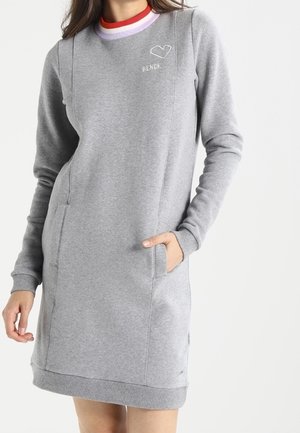 Femme portant une robe sweat-shirt grise à manches longues, longueur genou, avec poches latérales et rayures colorées sur le col, main dans la poche.
