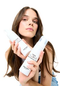 Białe butelki szamponu i odżywki trzymane przez osobę, z czarnym tekstem na butelkach. Tło jest jednolicie białe, co zwiększa widoczność produktu.