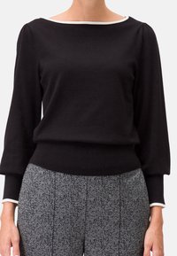 Schwarzer Strickpullover mit langen Ärmeln, weißem Kragenbesatz und geripptem Saum. Kombiniert mit grauen Hahnentrittmuster-Hosen.