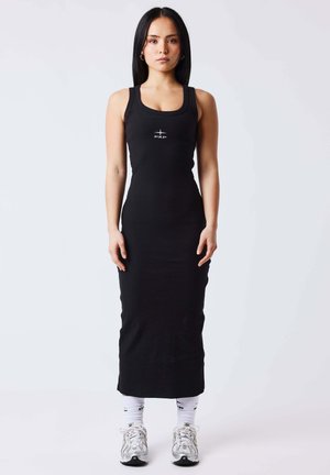 Mujer de pie, mirando hacia adelante, vistiendo un vestido ajustado negro sin mangas, calcetines blancos y zapatillas plateadas sobre un fondo liso.