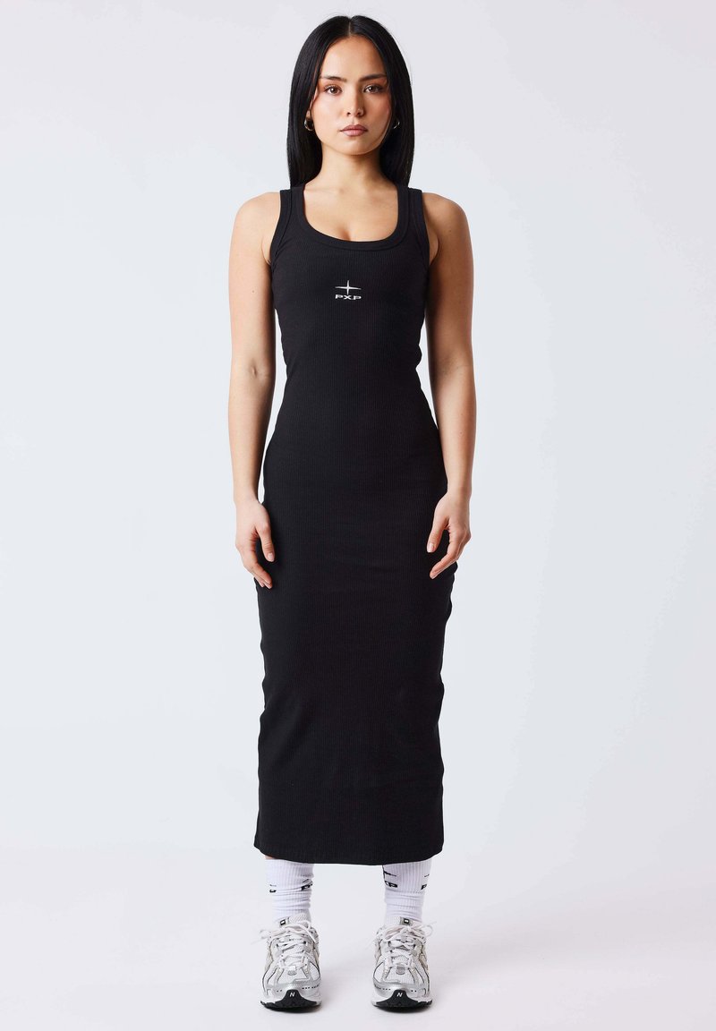 Femme debout de face portant une robe noire sans manches ajustée, des chaussettes blanches et des baskets argentées sur un fond uni.