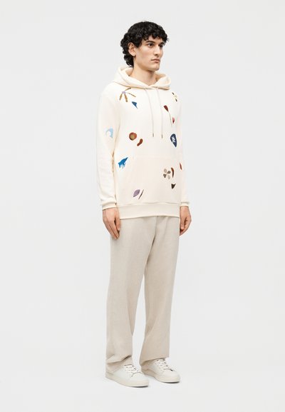 Mladý muž v krémovej mikine s viacfarebnými abstraktnými potlačami, béžových voľných nohaviciach a bielych teniskách, stojaci pred svetlým pozadím.