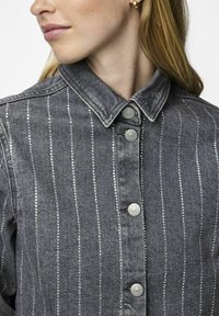 Giubbotto in denim grigio con strisce argento verticali, dotato di collo e bottoni a pressione in metallo. La texture appare robusta e strutturata.