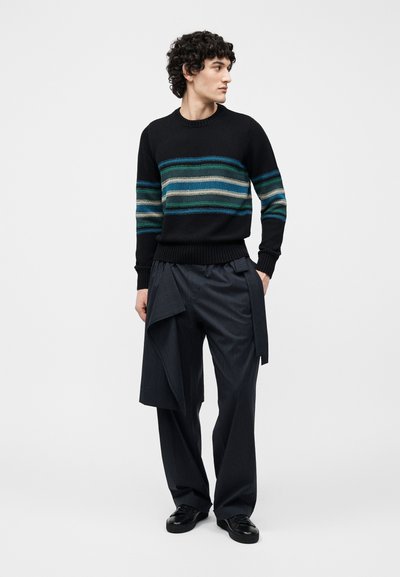 Homme aux cheveux bouclés portant un pull noir avec des rayures bleues et vertes, un pantalon large à fines rayures avec un détail en tissu, et des chaussures noires, debout et regardant vers la droite.