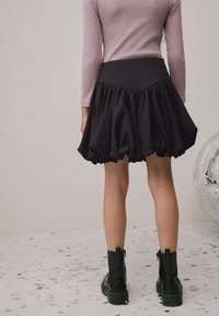 Gonna corta nera con orlo arricciato, tessuto strutturato e vita aderente; abbinata a stivaletti neri su una superficie scintillante.