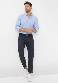 Chemise bleu clair à manches longues avec boutons, pantalon slim bleu marine foncé, et baskets blanches. Caractérisée par une poche poitrine unique et des manches retroussées.
