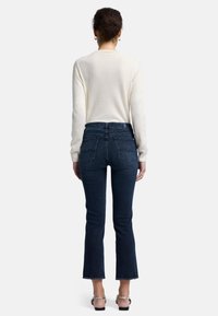 Un jean évasé bleu foncé avec une taille haute, un ourlet brut et des poches arrière. Porté avec un haut à manches longues de couleur claire.