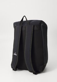 Svart Adidas ryggsäck med vadderade axelremmar, nätbakpanel, handtag upptill och gul sömnad mot en vit bakgrund.