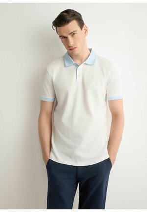 Młody mężczyzna z ciemnymi włosami, w białej koszulce polo z jasnoniebieskim kołnierzykiem i granatowych spodniach, stoi na białym tle.