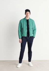 Veste matelassée verte avec fermeture éclair sur le devant, col rond, deux poches latérales, portée sur un haut noir et associée à un pantalon bleu marine et des baskets blanches.