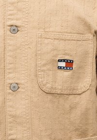 Μπεζ υφασμάτινη υφή με τσέπη στο στήθος που φέρει ένα πατς με το λογότυπο Tommy Jeans και δύο μεταλλικά κουμπιά με την ένδειξη Tommy Jeans.