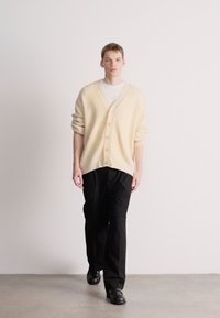 Krém színű, túlméretezett kardigán V-kivágással és gombos záródással, fekete nadrággal és fekete cipővel párosítva, egyszerű háttér előtt.