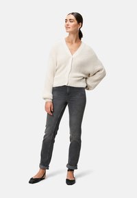 Cremfarbener Strickcardigan mit V-Ausschnitt, oversized Passform und gerippter Textur, kombiniert mit dunkelgrauen Slim-Fit-Jeans und schwarzen flachen Schuhen.