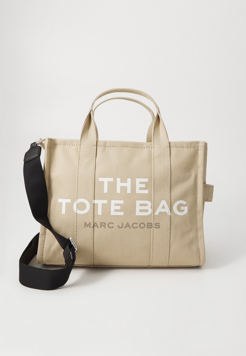 Μπεζ καμβάς τσάντα tote με διπλές επάνω λαβές, αφαιρούμενος μαύρος ιμάντας ώμου και μεγάλη λευκή επιγραφή που γράφει "Η ΤΣΑΝΤΑ TOTE MARC JACOBS."