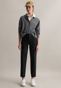 Grau gestrickter Pullover mit Knopfleiste, kombiniert mit hoch taillierter schwarzer Hose und weißen Turnschuhen, abgerundet mit einer beigefarbenen Mütze mit Logo.