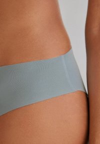 Lichtblauwe naadloze slip, met een gladde textuur en een brede tailleband, geplaatst tegen een huidskleurige achtergrond.