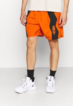 Homme portant un short de sport orange et noir avec un logo, des baskets montantes blanches Nike et des chaussettes noires, debout sur un sol uni.