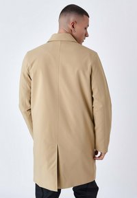 Abrigo beige con una textura suave y un diseño aerodinámico; cuenta con un cuello, abertura en la espalda y costuras reforzadas a lo largo de las costuras.