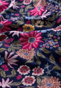 Le tissu floral présente un fond bleu marine foncé orné de fleurs vibrantes roses, jaunes et bleues, rehaussées de feuilles vertes et de détails complexes.