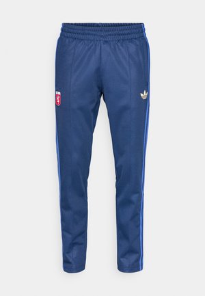 Jūriniai treniruočių kelnės, pagamintos iš lygaus audinio, su elastišku juosmeniu, mėlynomis šoninėmis juostelėmis ir auksiniu Adidas logotipu kairėje klubo pusėje.