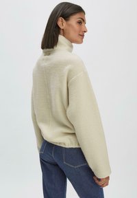 Helle beigefarbene Fleece-Sweatshirt mit hohem Kragen, oversized Schnitt und überschnittenen Schultern, kombiniert mit dunklen Jeans.