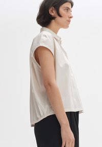 Short-sleeve beige blouse in lightweight fabric, featuring a button closure and gathered hem, paired with black pants. Smooth texture.