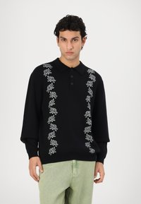 Zwarte sweatshirt met een kraag, voorzien van witte rozenborduursels langs het midden. De mouwen zijn lang en de stof lijkt zacht.
