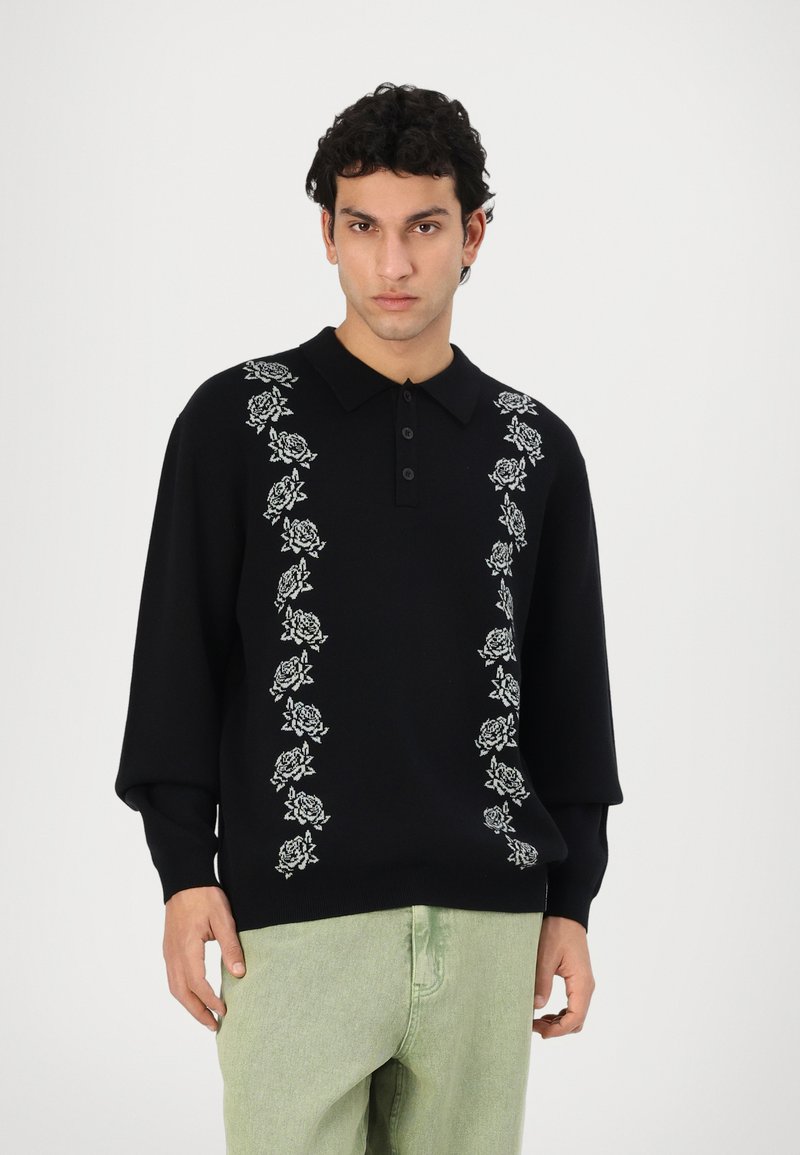 Zwarte sweatshirt met een kraag, voorzien van witte rozenborduursels langs het midden. De mouwen zijn lang en de stof lijkt zacht.