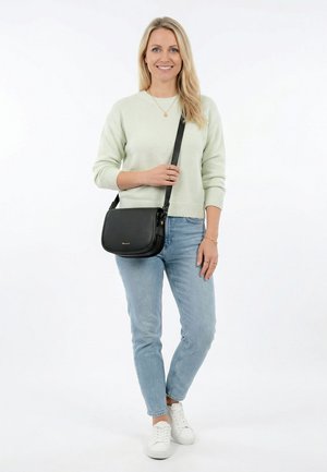 Mujer rubia sonriendo, vestida con un suéter ligero, jeans azules, zapatillas blancas y llevando un bolso negro de bandolera sobre el hombro.