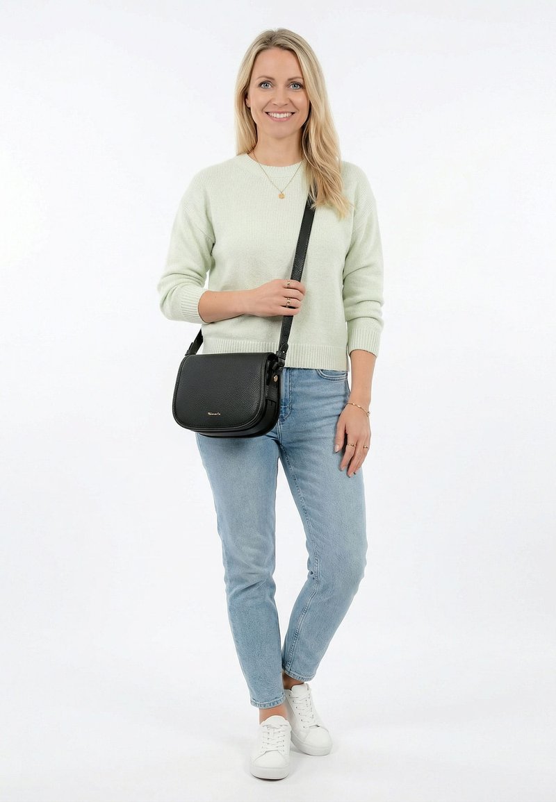 Femme blonde souriante, portant un pull léger, un jean bleu, des baskets blanches et un sac bandoulière noir sur l'épaule.