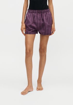 Person trägt locker sitzende Boxershorts in Lila und dunklen Streifen sowie ein schwarzes Tanktop, steht barfuß vor weißem Hintergrund.