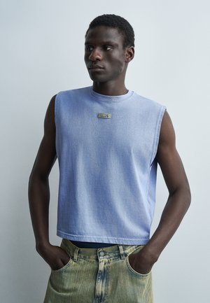 Νέος άνδρας με κοντά μαλλιά, ντυμένος με αμάνικο γαλάζιο πάνω μέρος και ξεβαμμένο κίτρινο τζιν, στέκεται με τα χέρια στις τσέπες μπροστά σε ένα απλό φόντο.