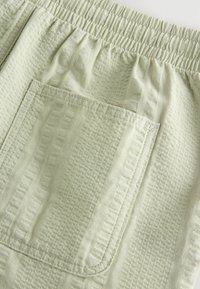 Tissu vert clair texturé avec une ceinture élastique et une grande poche rectangulaire cousue à l'arrière.
