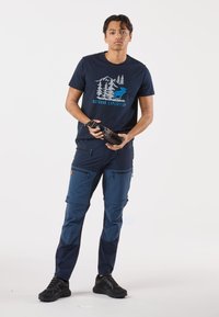 Mörkblå t-shirt med utomhusmotiv; mörkblå cargobyxor med flera blixtlåsfickor; svarta träningsskor; håller i en svart vattenflaska.