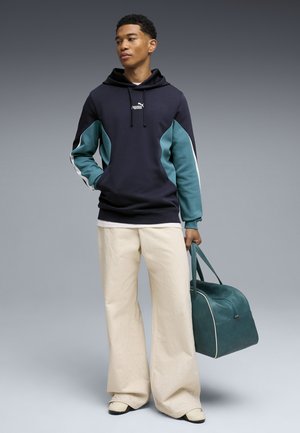 Jonge man staand in beige wijde broekspijpen, donker hoodie met blauwe mouwen, houdend een groene reistas tegen een grijze achtergrond.