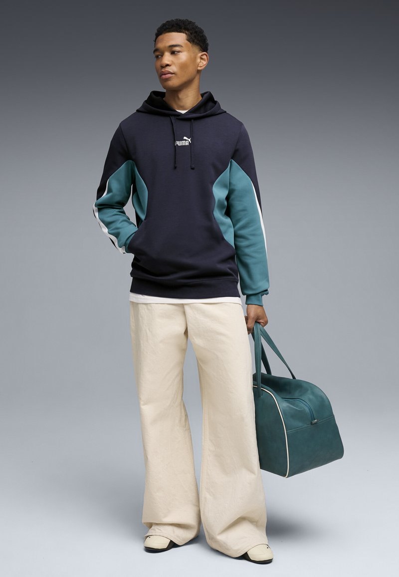 Jeune homme debout en pantalon beige à jambes larges, sweat à capuche sombre avec manches bleues, tenant un sac de sport bleu sarcelle sur un fond gris.