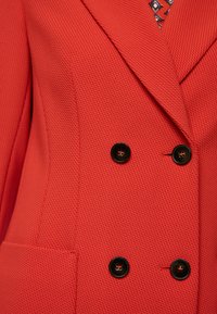 Roter strukturierter Blazer mit Reverskragen, zweireihigem Design und schwarzen Knöpfen mit roten Ziernähten.