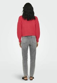 Afgeknotte roze gebreide trui met brede mouwen, gecombineerd met lichtgrijze rechte jeans; model draagt zwarte sandalen. Zicht van achteren.