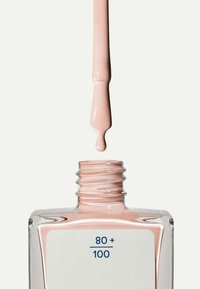 Nagellack i en klar glasflaska med fyrkantig form, i en mjuk rosa färg. Penseln är doppad i nagellacket och droppar ovanför öppningen.