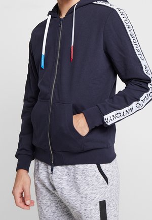 Mężczyzna ubrany w granatową bluzę na zamek z białymi paskami z logo na rękawach i sznurkami, w połączeniu z jasnoszarymi spodniami dresowymi z czarnymi detalami.